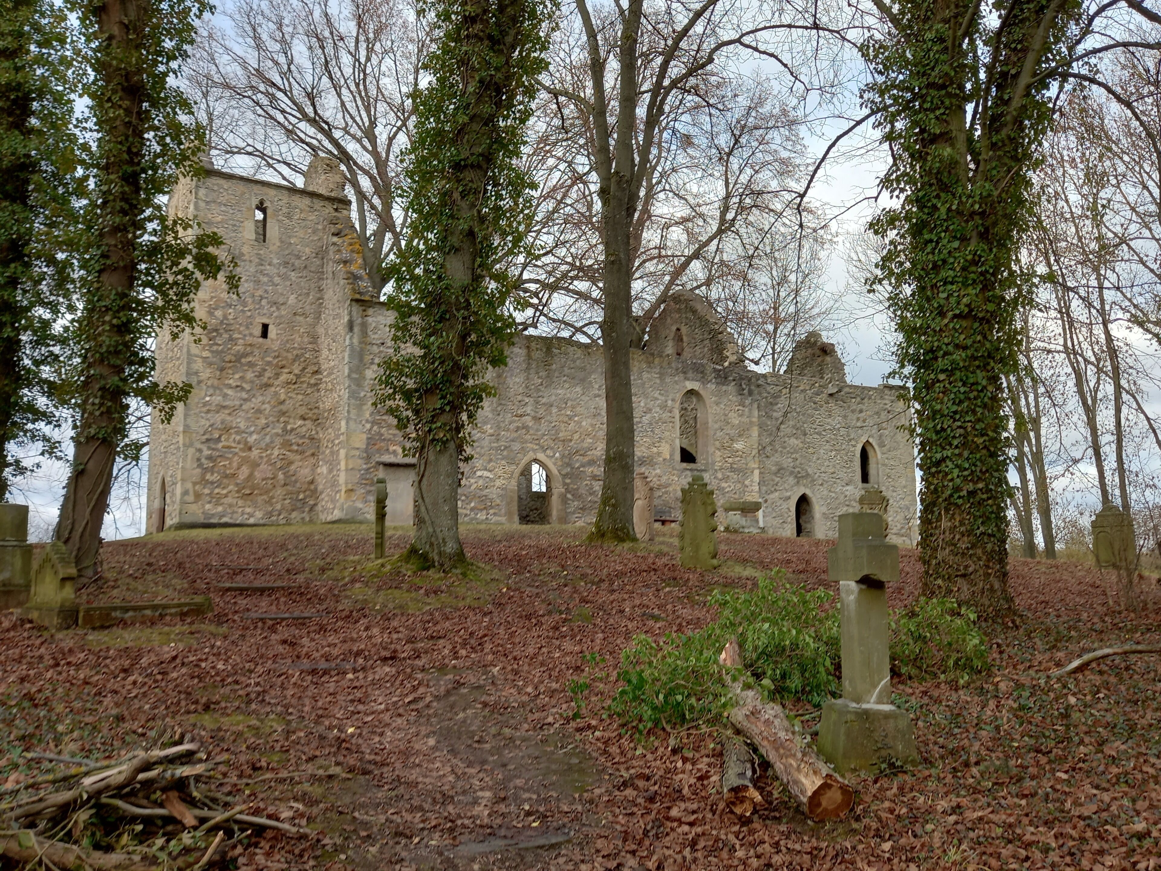Ein altes Steinhaus mit Rundbogenfenstern und einem zentralen Eingang, umgeben von Bäumen und einem Friedhof.