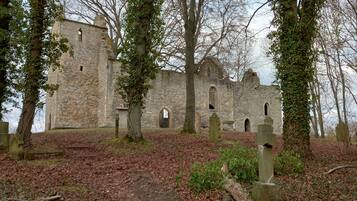 Ein altes Steinhaus mit Rundbogenfenstern und einem zentralen Eingang, umgeben von Bäumen und einem Friedhof.