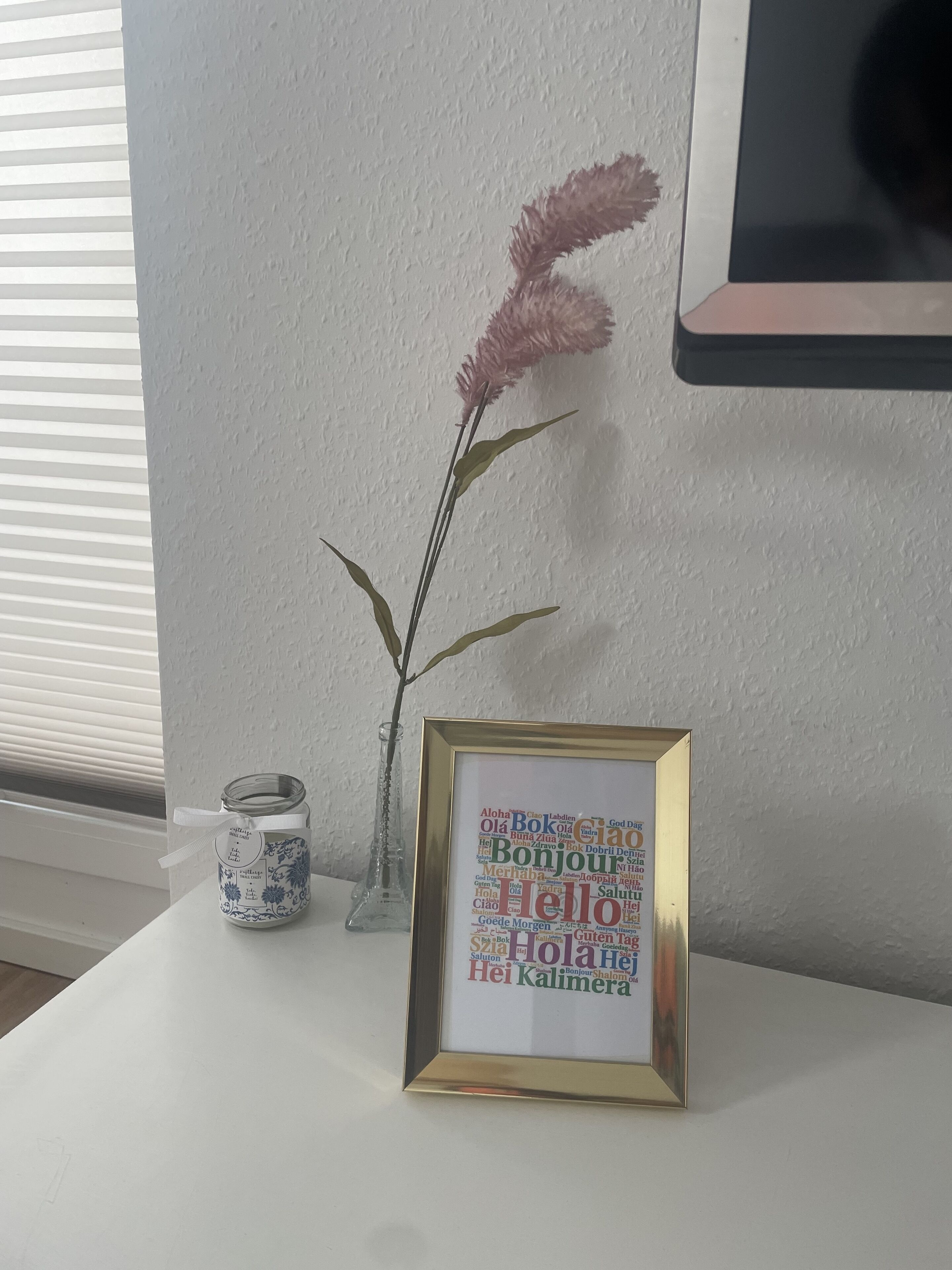 A framed word cloud with greetings in multiple languages, a small vase with pink flowers, and a decorative jar on a white table.