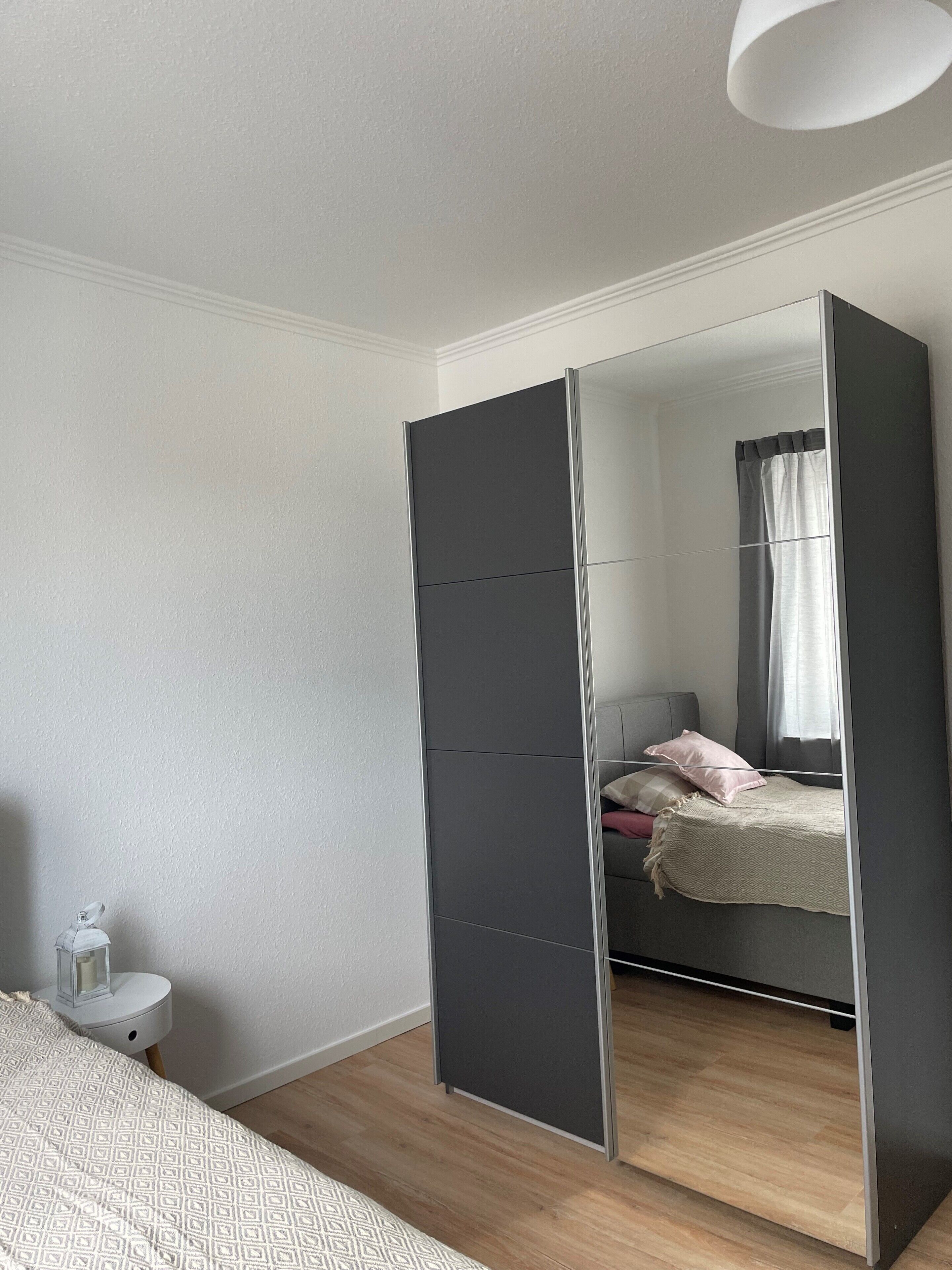 A bedroom with a large wardrobe, a bed with a patterned bedspread, a bedside table with a candle, and a window with curtains.