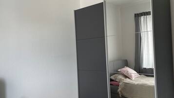 A bedroom with a large wardrobe, a bed with a patterned bedspread, a bedside table with a candle, and a window with curtains.