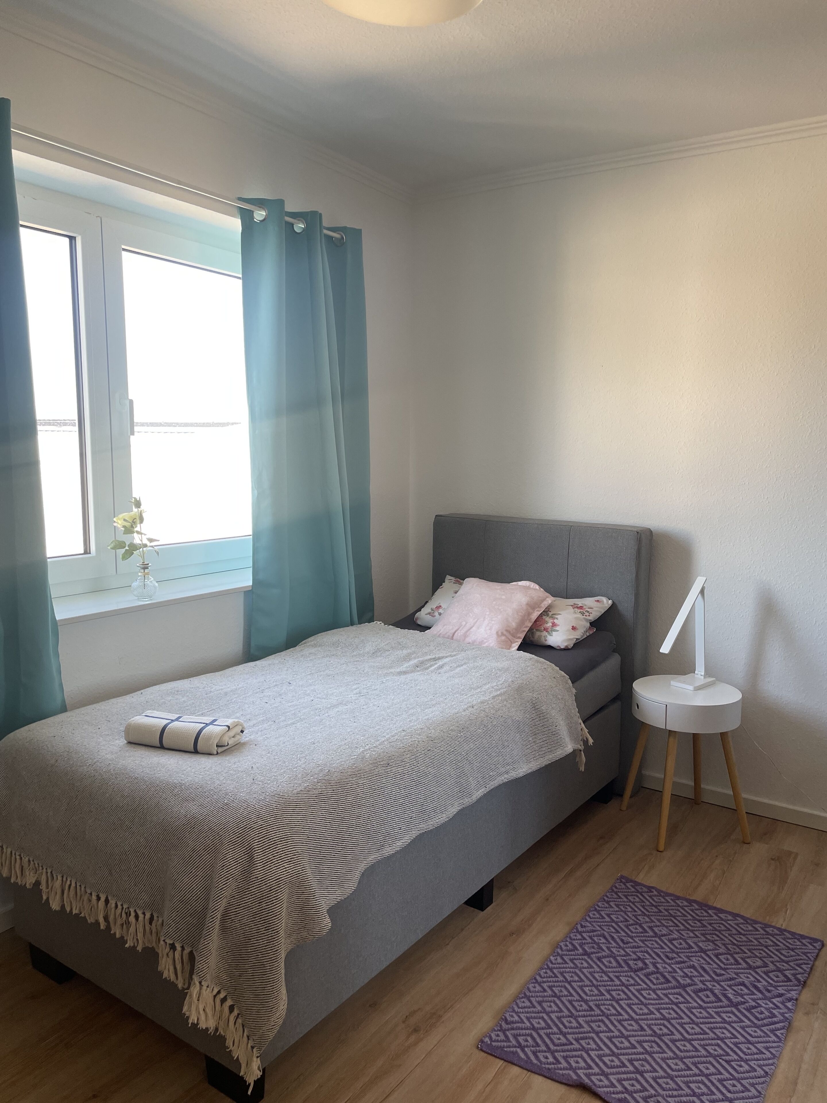 Ein ordentlich eingerichtetes Schlafzimmer mit Bett, Nachttisch, einem Fenster mit Vorhängen und einem kleinen Teppich.