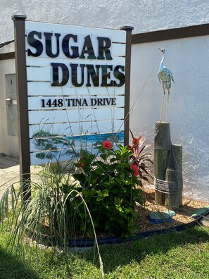Placa de 'Sugar Dunes' no número 1448 da Tina Drive, com uma escultura de pássaro e vegetação ao redor.