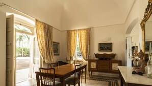 Un comedor con una mesa de madera, sillas y un armario. Hay una ventana con cortinas, un espejo y una estatua sobre la mesa.
