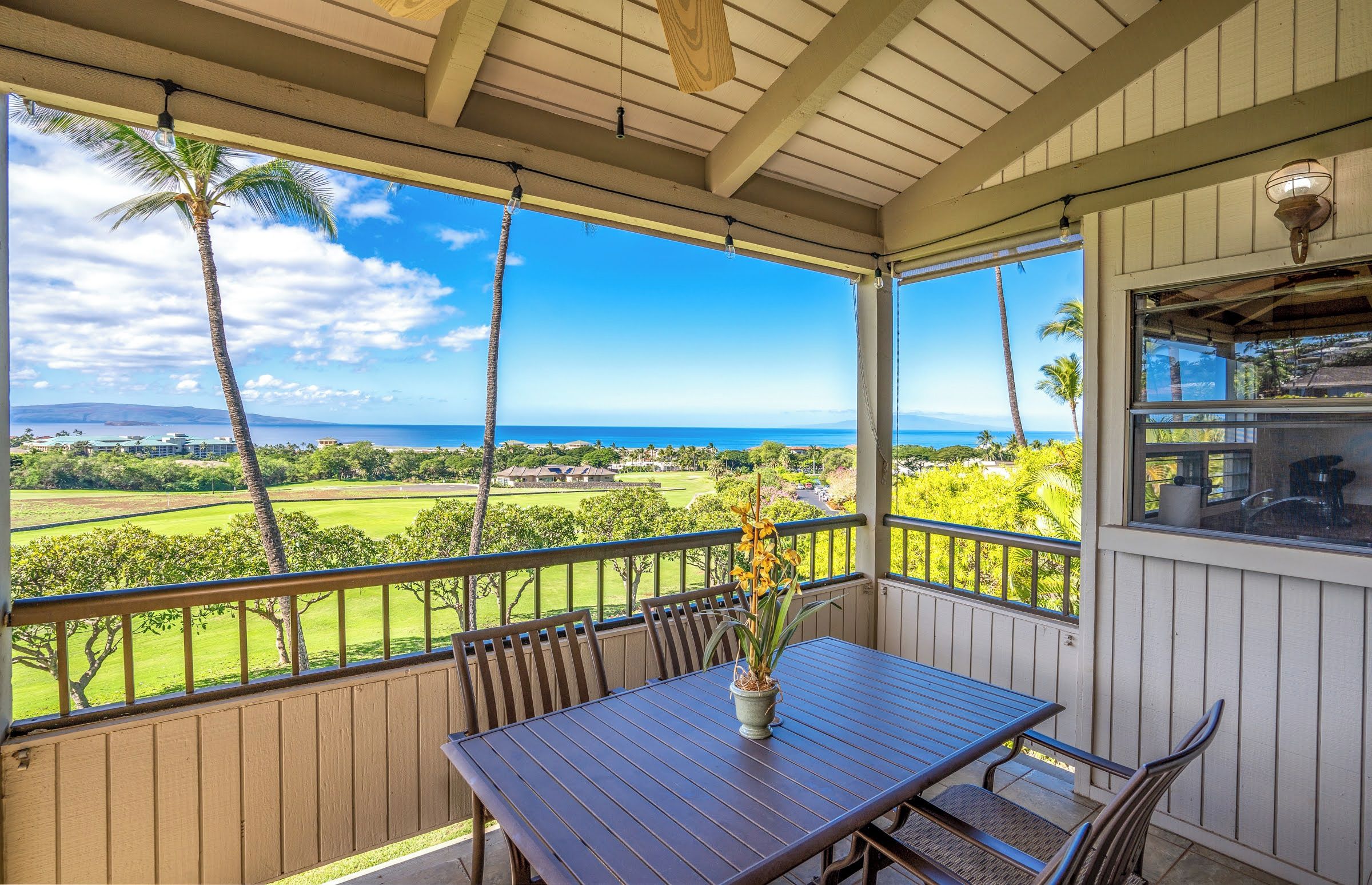A covered outdoor area with a wooden table and chairs, a potted plant, and a view of a golf course and ocean.