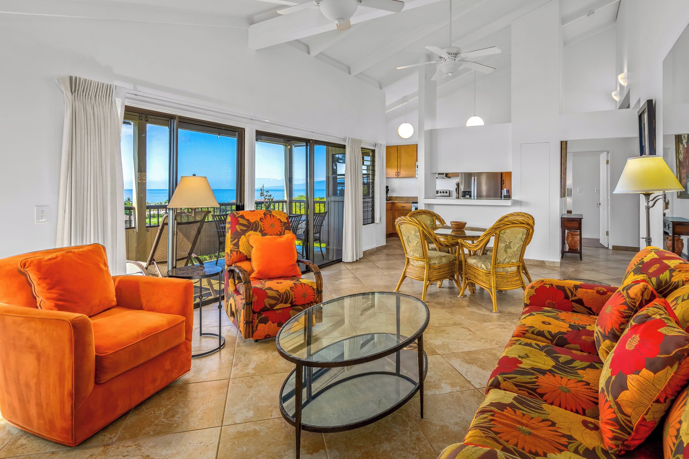 A spacious living area with a sofa, armchairs, and a glass coffee table. A kitchen is visible in the background, and large windows offer a view of the outdoors.