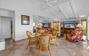 A spacious living area with a dining table surrounded by wicker chairs, a large flat-screen TV, and a comfortable sofa.