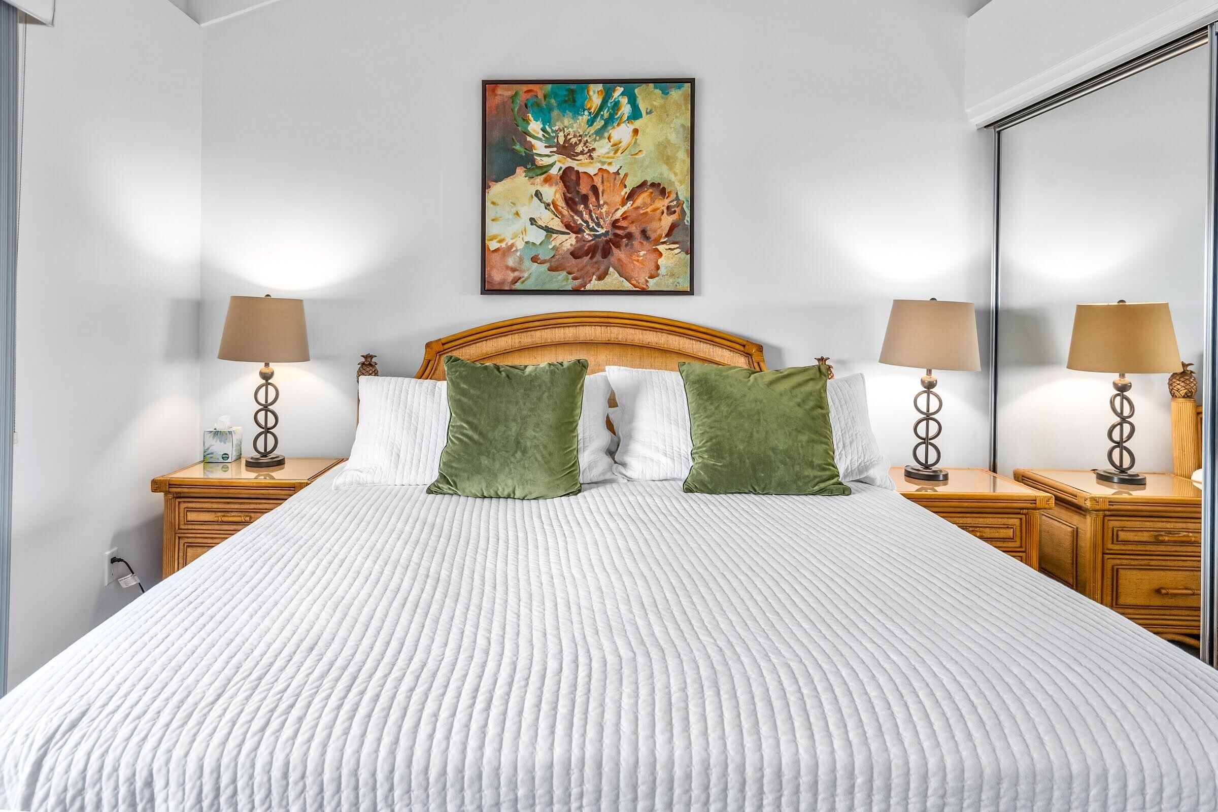 A neatly made bed with a white quilt, two green pillows, and a wooden headboard. There are two bedside tables with lamps and a painting above the bed.