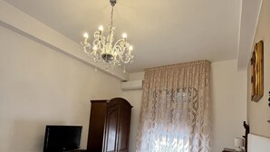 A hotel room with a bed, a television, a wooden wardrobe, a chandelier, and patterned curtains.