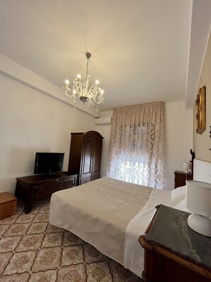 Quarto de hotel com cama, televisão, guarda-roupa de madeira, candelabro e cortinas estampadas.