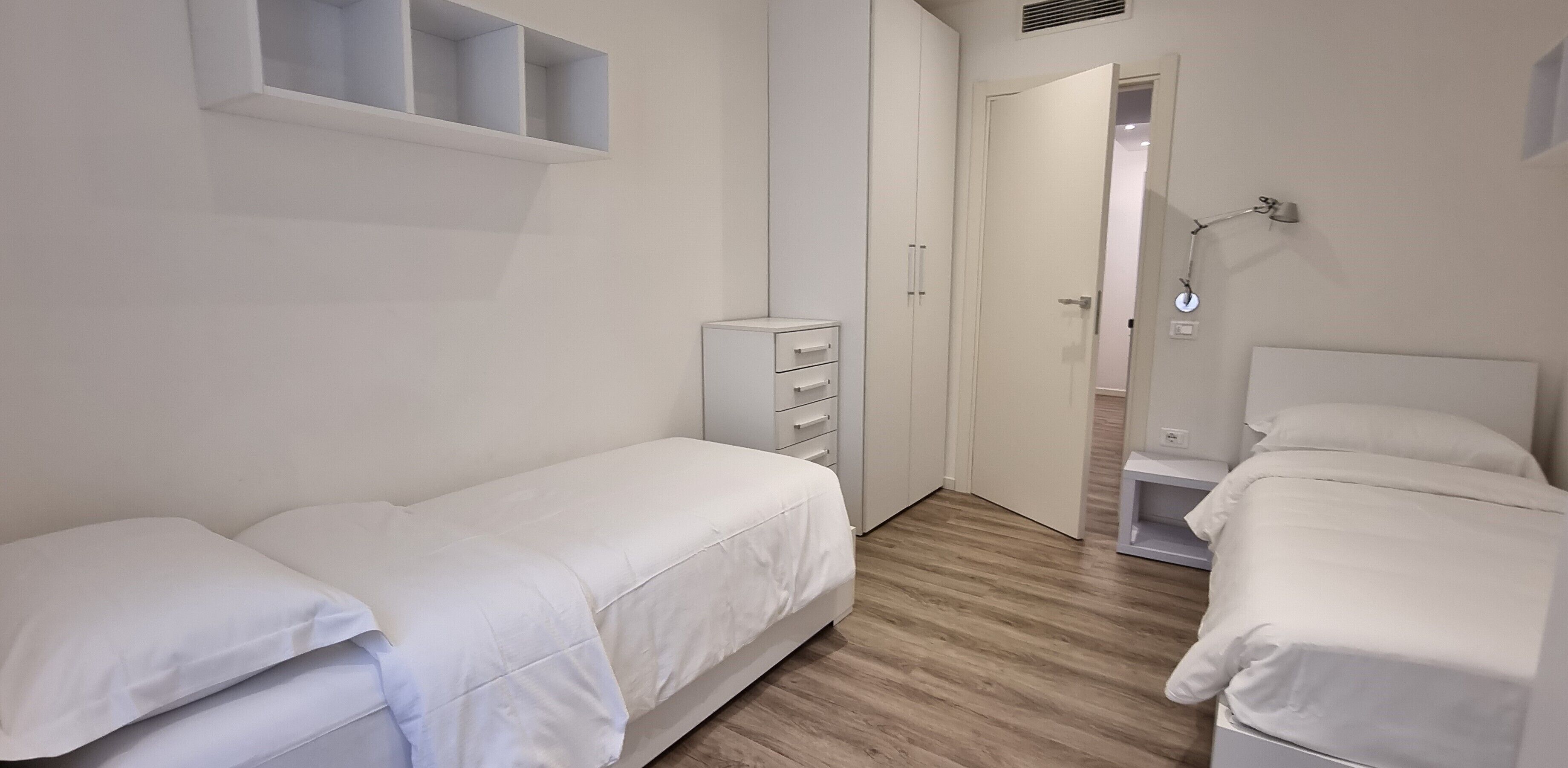 Un dormitorio con dos camas individuales, un armario, una mesita de noche y un estante sobre la cama.