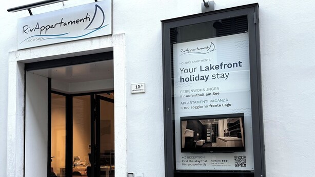 Ein Hotel Eingang mit einem Schild, auf dem 'RivApartmenti' steht, und einer Fensterwerbeanzeige für 'Your Lakefront holiday stay'.