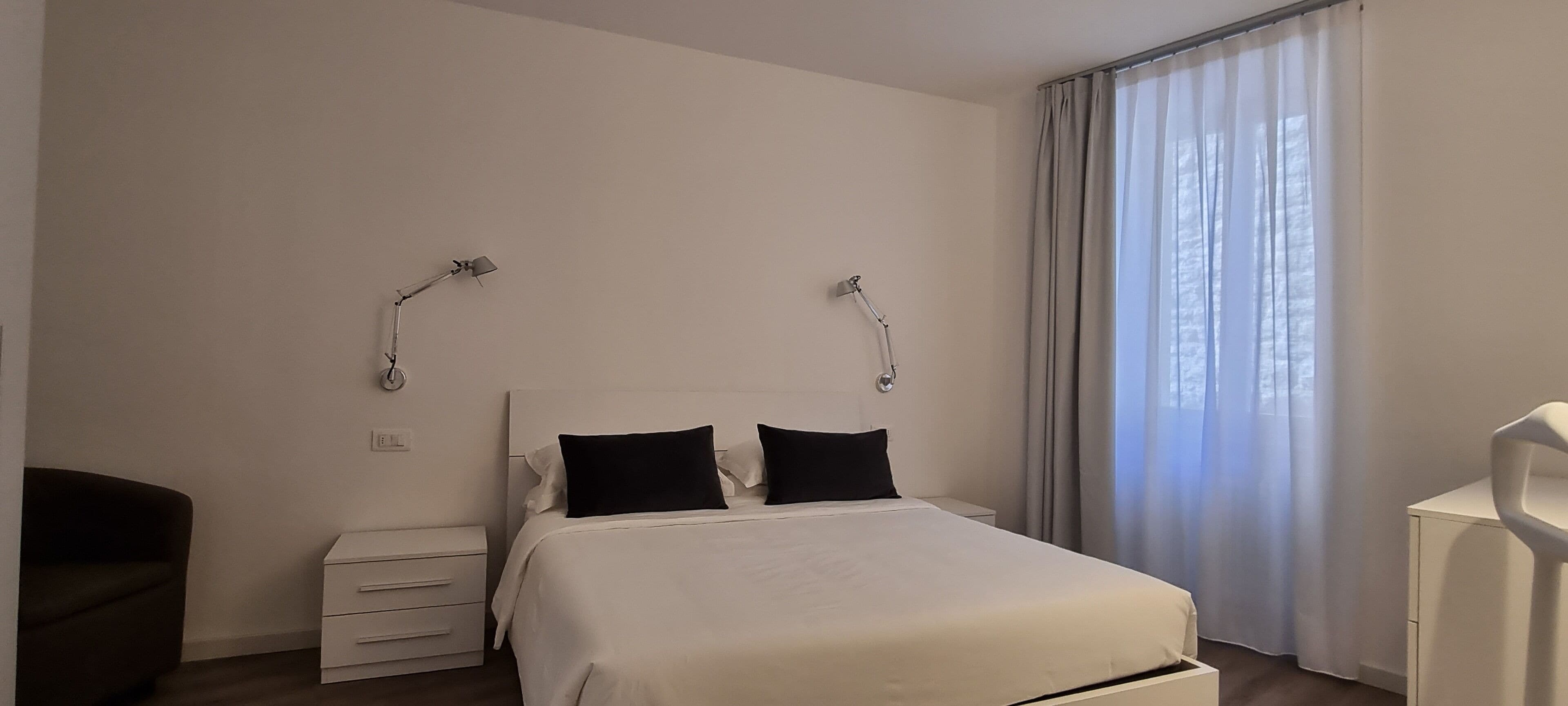 Hotelkamer met een bed, twee bedlampjes, een nachtkastje, een stoel en een raam met gordijnen.