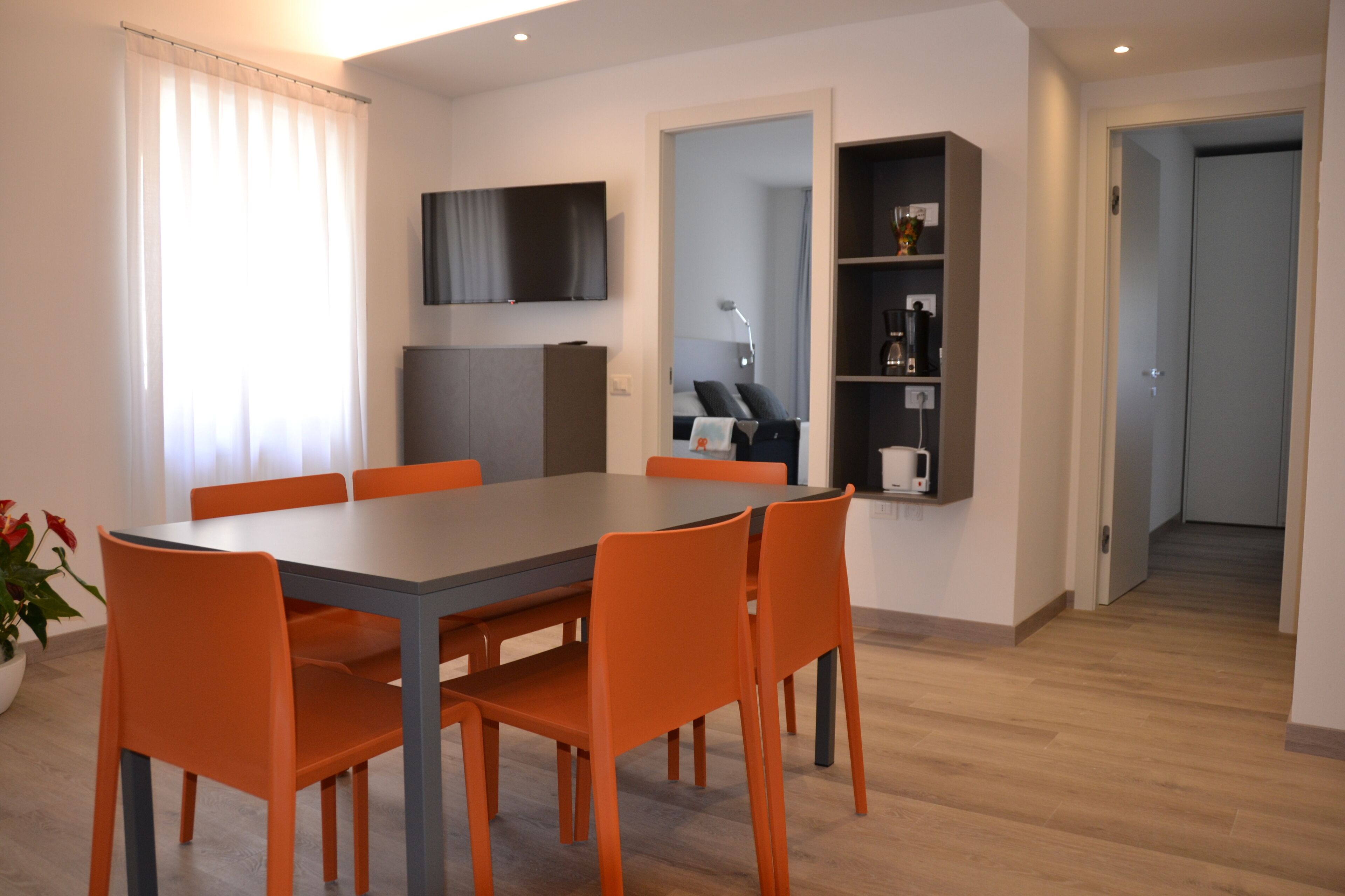 Et moderne spiseområde med et rektangulært bord og seks orange stole. Et fladskærms-tv er monteret på væggen over bordet.