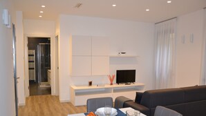 Ein modernes Wohnzimmer mit Essbereich, einer Couch, einem Fernseher auf einem weißen Schrank und einer Glastür zum Badezimmer.
