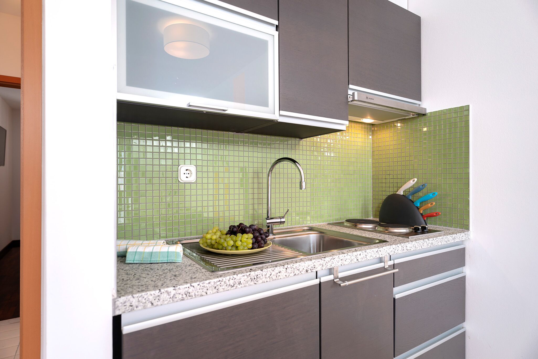 Uma cozinha com painel de azulejos verdes, uma pia de aço inoxidável e bancada com uma fruteira.