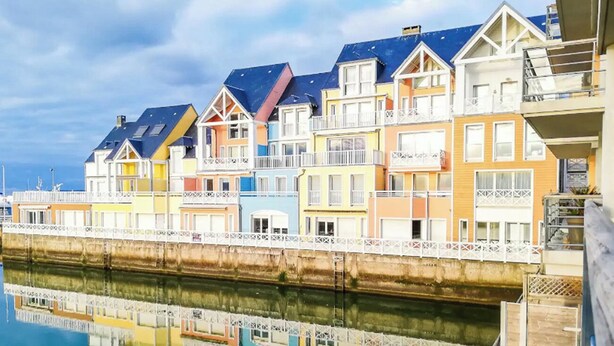 Eine Reihe bunter Häuser entlang einer Uferpromenade, deren Spiegelung sich klar im Wasser zeigt.