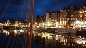 Ein beleuchteter Yachthafen bei Nacht, mit vertäuten Booten, darunter ein markantes Segelboot im Vordergrund, und Häusern entlang der Uferpromenade.