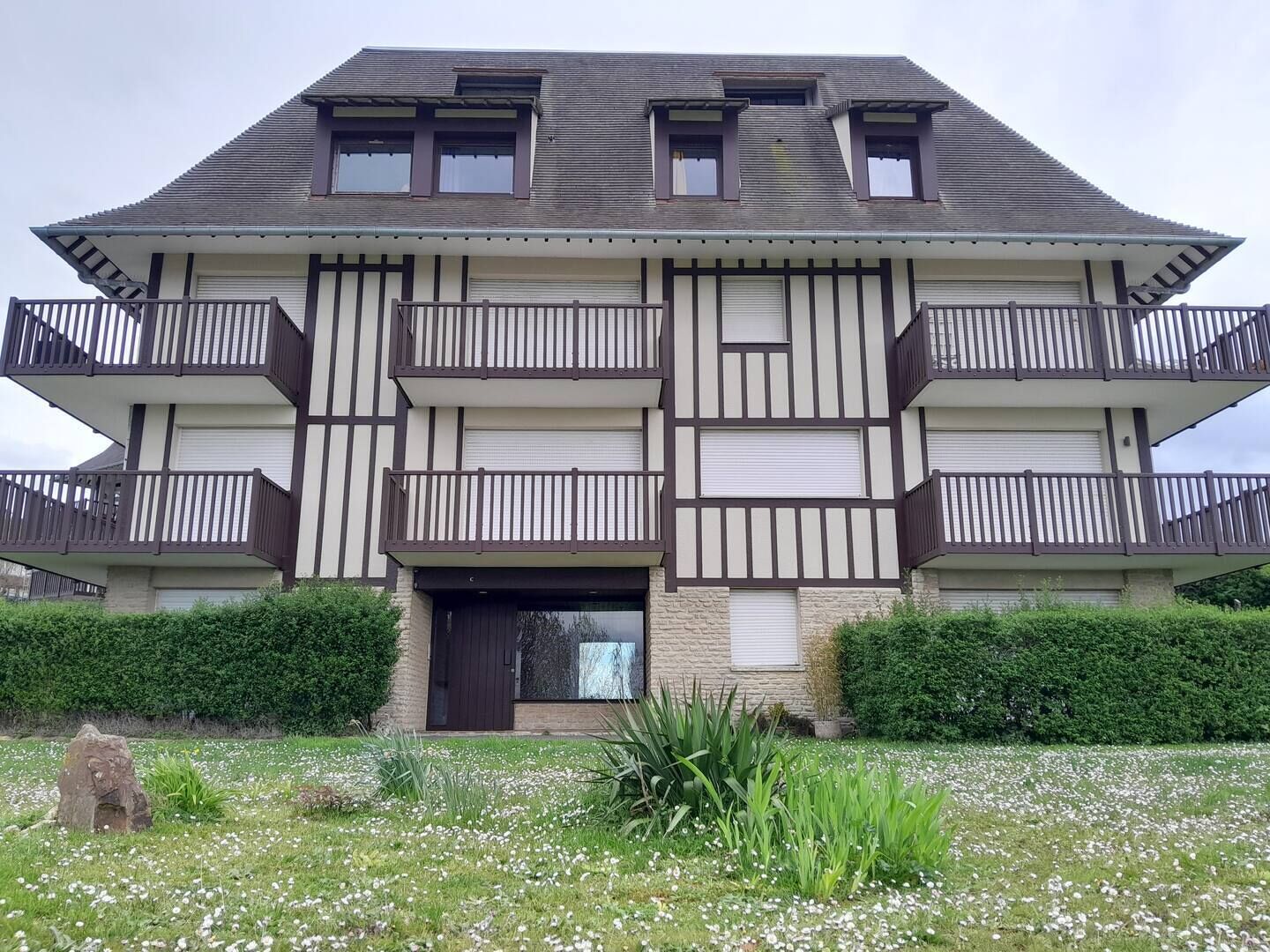 A multi-story residential building with balconies, a front entrance, and a garden area with flowers.