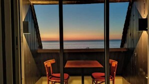 Ein Zimmer mit Holzbalkendecke, zwei roten Stühlen, einem Holztisch und einem großen Fenster mit Blick auf den Ozean bei Sonnenuntergang.
