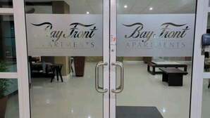 A glass door with the text "Bay Front Apartments" and a view of an interior space with a reception desk and seating area. - Ornella Villa - 2-br Apartment - Pool - Parking (Portmore)