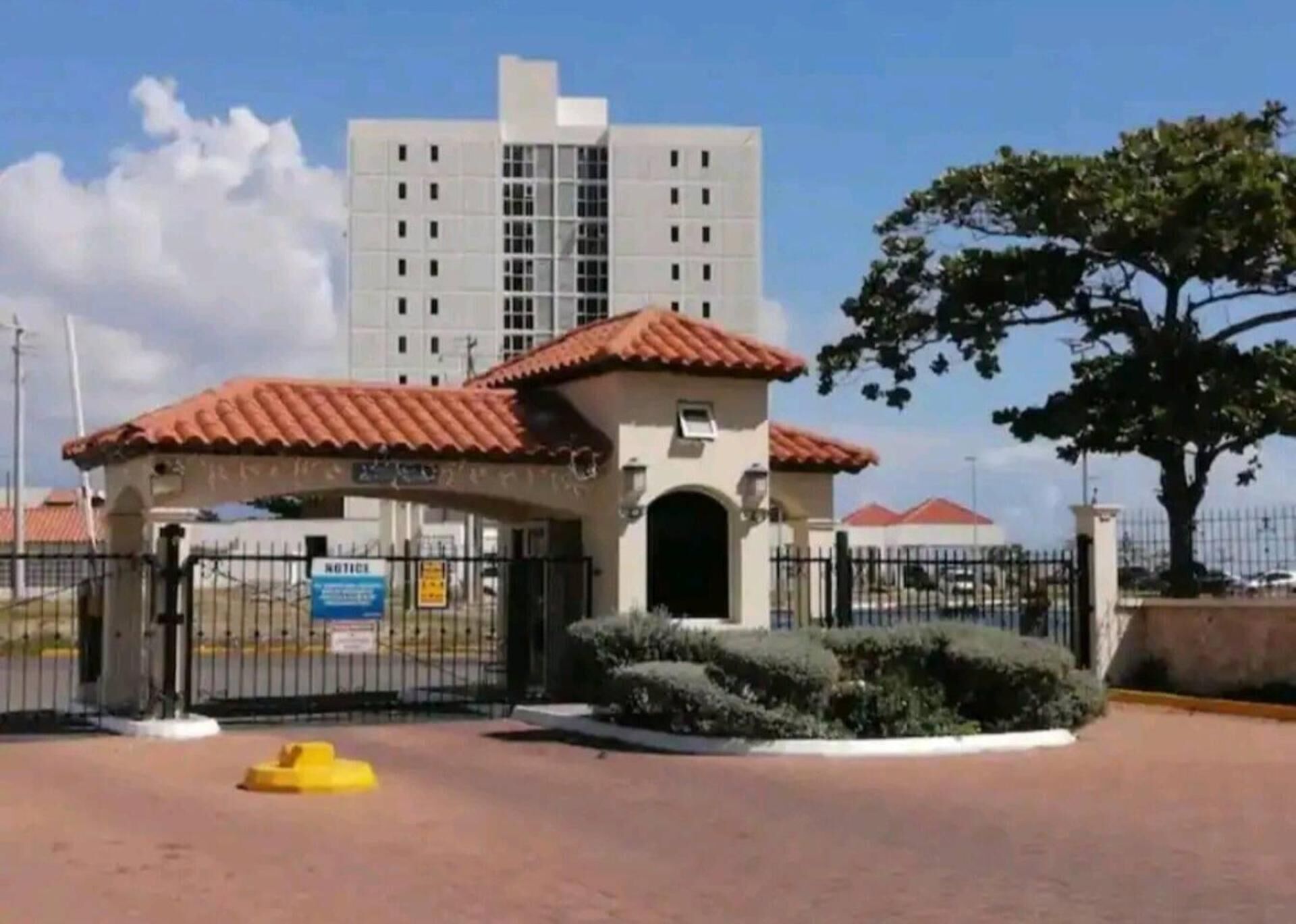 A gated entrance with a red-tiled roof, a small building, and a tall white building in the background.