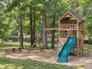 Un parque infantil con un tobogán de madera y columpios, rodeado de árboles y un lago.