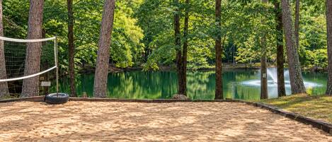 A sandy volleyball court with a net, surrounded by trees and a calm lake.