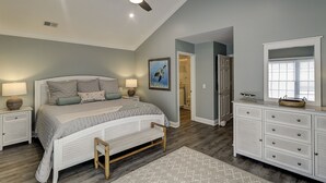 Un dormitorio con cama, mesitas de noche, cómoda, ventilador de techo, un cuadro y una puerta que da a otra habitación.