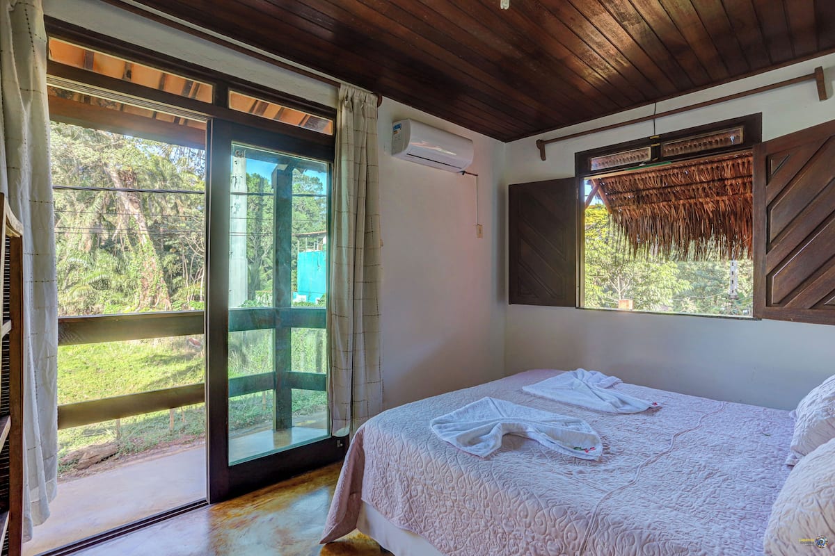 Quarto com uma janela grande, uma cama com colcha rosa e um aparelho de ar condicionado na parede.