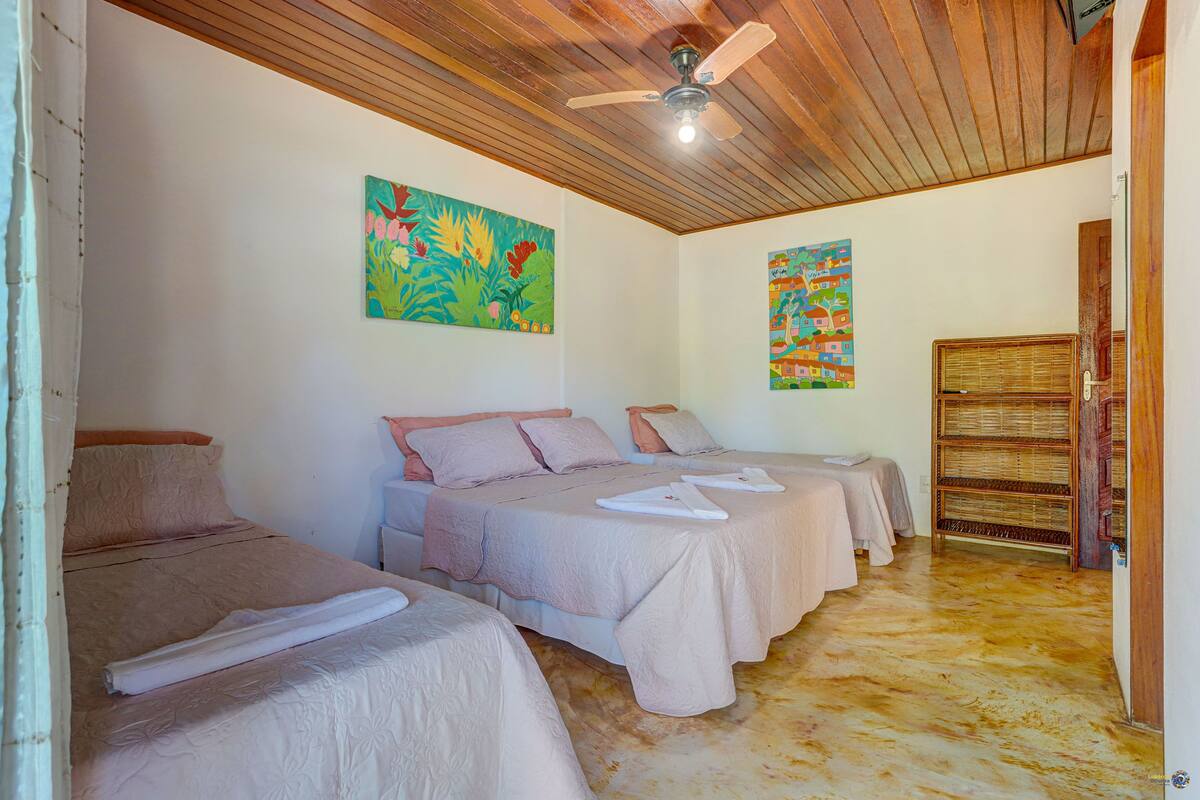 Quarto de hotel com três camas, teto de madeira e duas pinturas coloridas nas paredes.