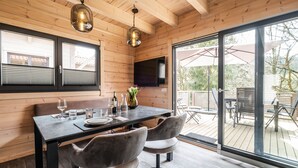 A wooden interior with a dining table set for two, a flat-screen TV mounted on the wall, and a sliding glass door leading to an outdoor patio. - Bergwaldchalets Wellness (Wernigerode)