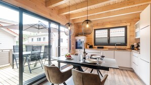 A modern kitchen with wooden beams, a dining area with a glass table and chairs, and a view of the outside. - Bergwaldchalets Wellness (Wernigerode)