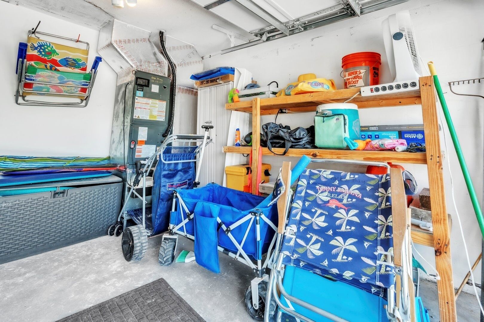 A compact, well-organized room with a bed, a small desk, and a chair. There are shelves with various items, including a fan, a bucket, and some bags.
