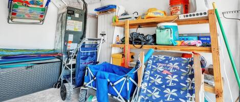 A compact, well-organized room with a bed, a small desk, and a chair. There are shelves with various items, including a fan, a bucket, and some bags.