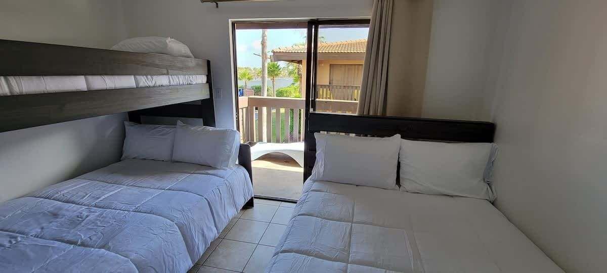 Una habitación con dos camas, cada una con ropa de cama blanca y cabeceros. Cuenta con un balcón que ofrece vistas a zonas verdes y un edificio.