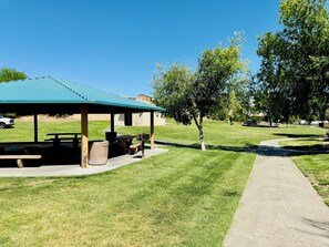 Un espace pique-nique abrité, équipé de bancs et d’un barbecue, entouré de pelouse et d’arbres bien entretenus.