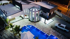 Uma casa moderna de dois andares com piscina, um alpendre coberto e um carro branco estacionado perto.