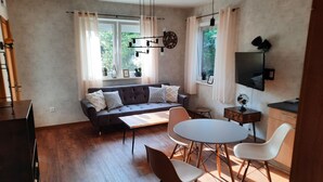 Ein modernes Wohnzimmer mit einer Couch, Esstisch und Stühlen. Es gibt Fenster mit Vorhängen, eine Deckenleuchte und ein flachbildfernseher an der Wand.