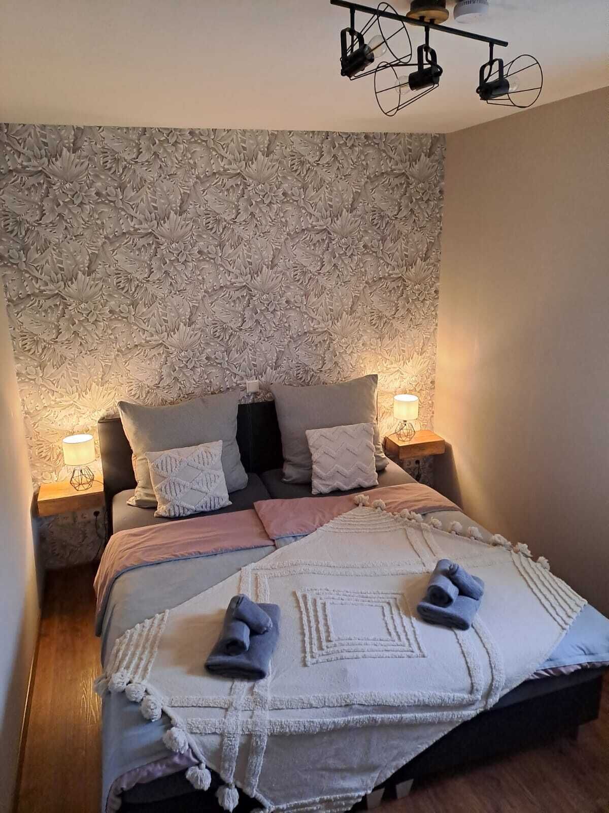 Ett sovrum med en blommig tapet, en säng med många kuddar, två sänglampor och en taklampa.
