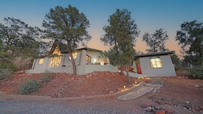 A modern two-story house with a stone foundation, surrounded by trees and a gravel driveway.