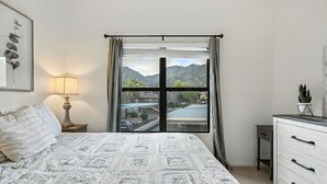 Un dormitorio con cama, mesita de noche con lámpara, cómoda y ventana con vistas a montañas y al paisaje urbano.