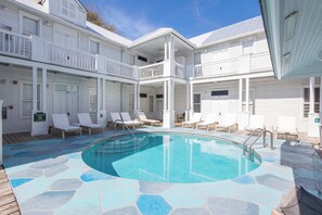 A pool area with a stone deck, white lounge chairs, and a two-story building with balconies.