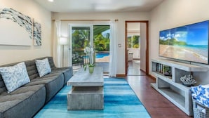 A modern living room with a grey sofa, a coffee table, and a TV on a stand. - Hale Ohia: Secluded Home w/ Private Pool, Ocean & Mountain Views, BBQ (Princeville)