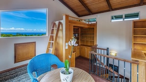 A room with a large window overlooking a beach, a wooden staircase, and a bookshelf. - Hale Ohia: Secluded Home w/ Private Pool, Ocean & Mountain Views, BBQ (Princeville)