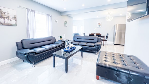 A modern living room with a sofa, ottoman, and coffee table, a dining area with chairs, and a kitchen with a refrigerator. - The Cedar Chateau (Niagara Falls)