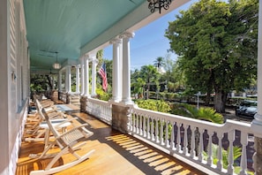 A covered porch with white columns and a railing, wooden rocking chairs, and a view of a landscaped garden with palm trees. - The Curry Poolside | Ground  Floor King (Key West)