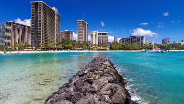 A coastal cityscape with high-rise buildings, a clear blue sky, and a rocky shoreline.