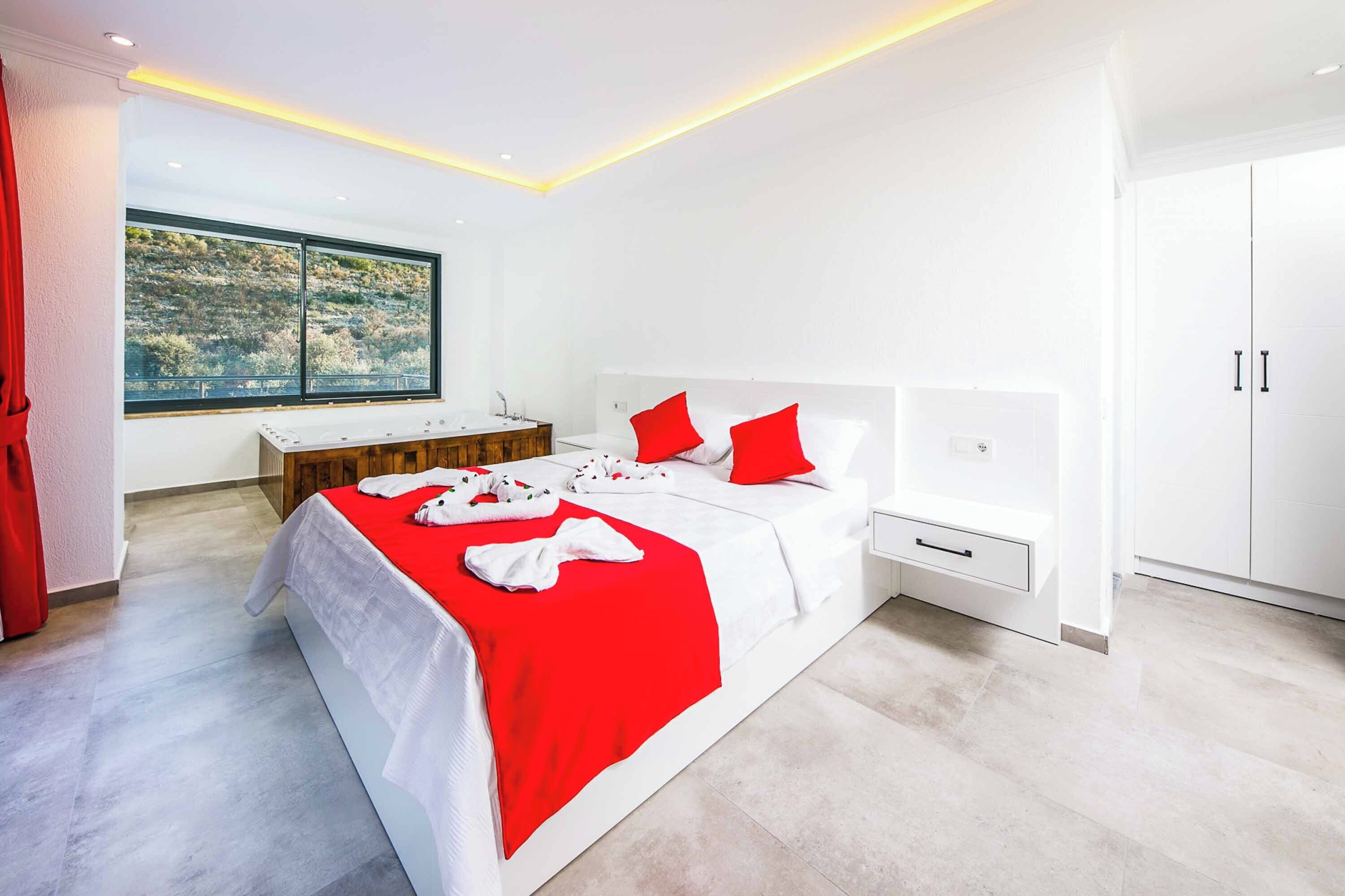 A modern bedroom with a large bed, white walls, a wooden bench, a large window with a view, and a white wardrobe.
