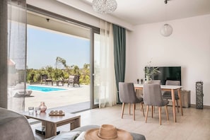 Ein modernes Wohnzimmer mit einer großen Schiebeglastür, die zu einem Poolbereich führt, einem Esstisch mit Stühlen und einem Flachbildfernseher.